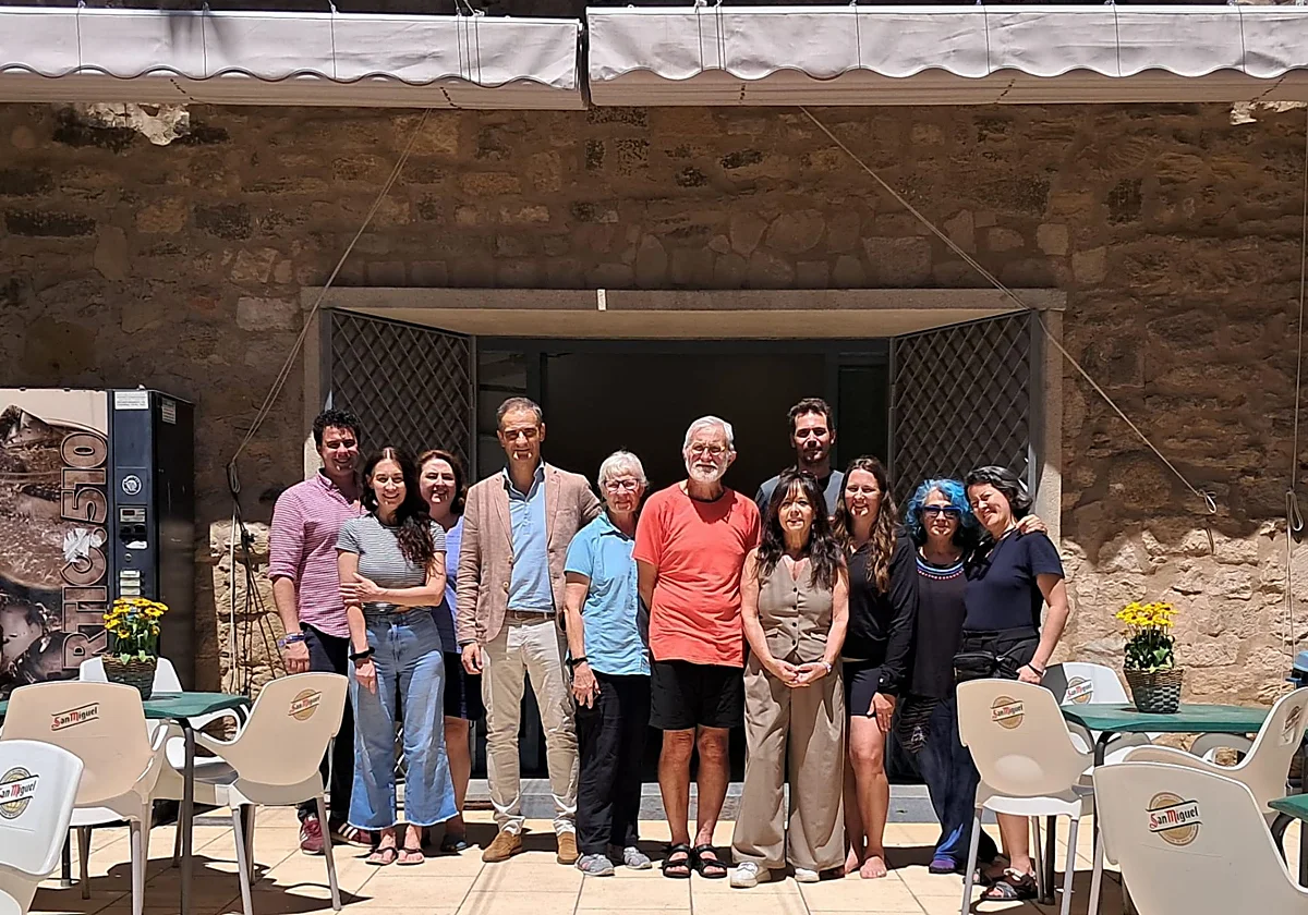 Group of twelve people posing together outside a stone building on a sunny terrace with tables and chairs. (Group photo on a café terrace)