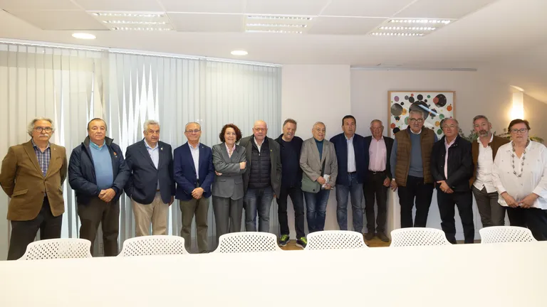 Group of adults standing in a conference room behind a long white table, posing for a group photo; everyone is dressed in business casual or formal attire, with a glass partition and framed artwork in the background.