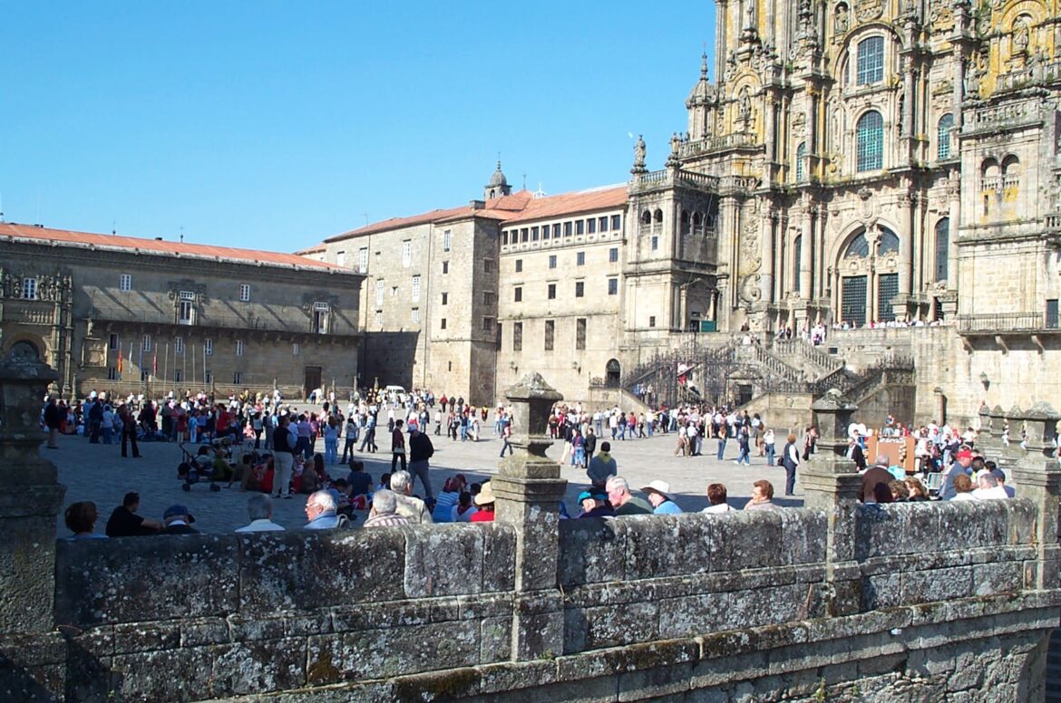 Crowded plaza in front of a grand, ornate cathedral with stone steps and a blue sky above. People are walking and sitting around the square.