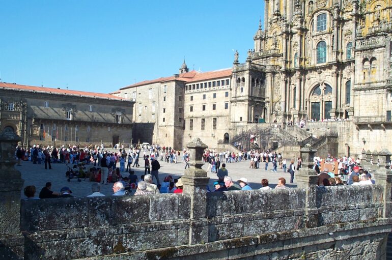 Crowded plaza in front of a grand, ornate cathedral with stone steps and a blue sky above. People are walking and sitting around the square.