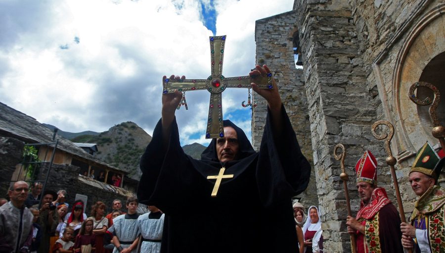 A priest in a black robe lifts a large ornate cross above a crowd outside a stone church, with onlookers and clergy nearby.