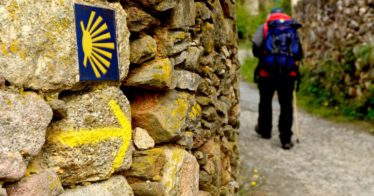 Stone wall with a blue tile showing a yellow scallop shell trail marker, and a hiker with a backpack walking along a gravel path ahead.