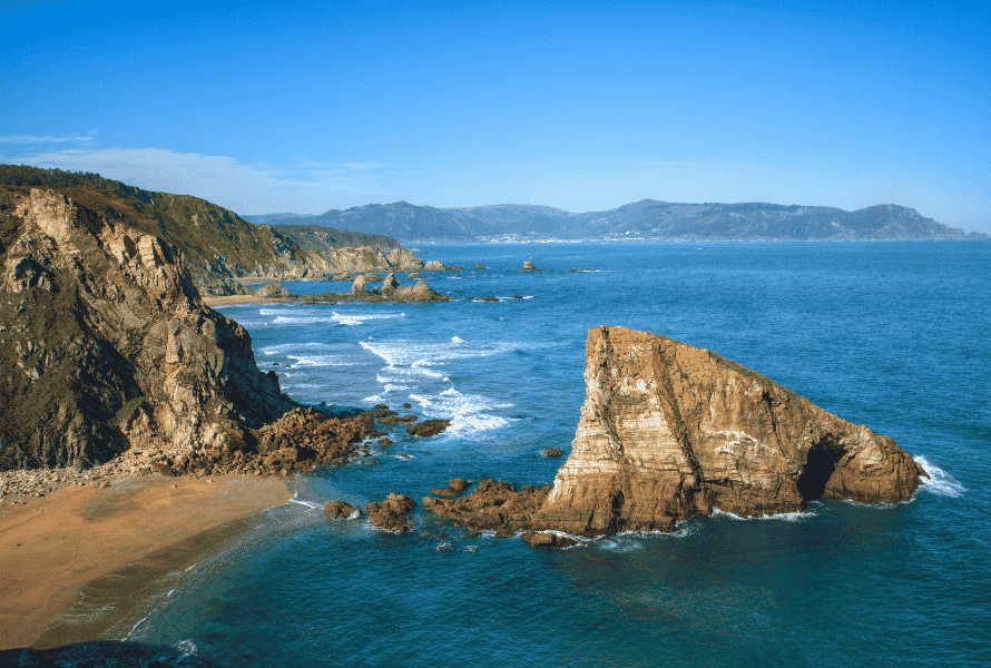 Rugged coastal scene with large rock formation rising from turquoise water and rocky cliffs along a sandy shore under a clear blue sky