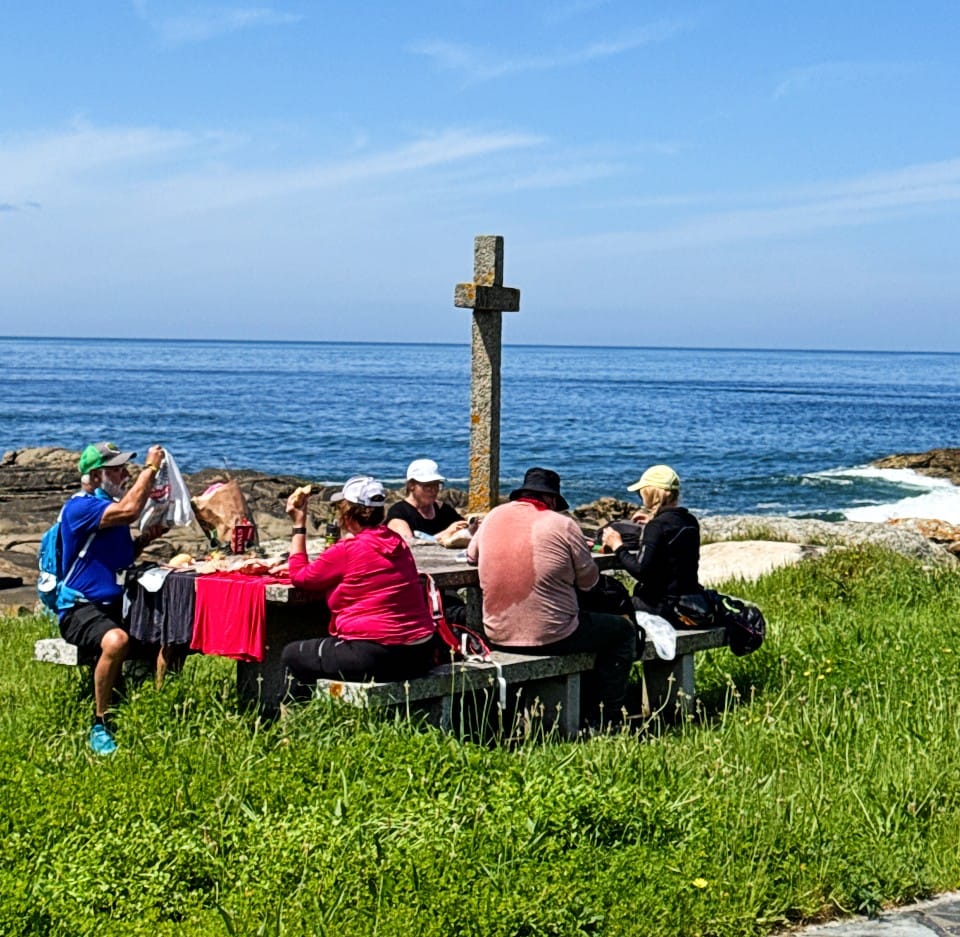 Group of people picnicking at a coastal stone table with a cross monument, ocean visible in the background.