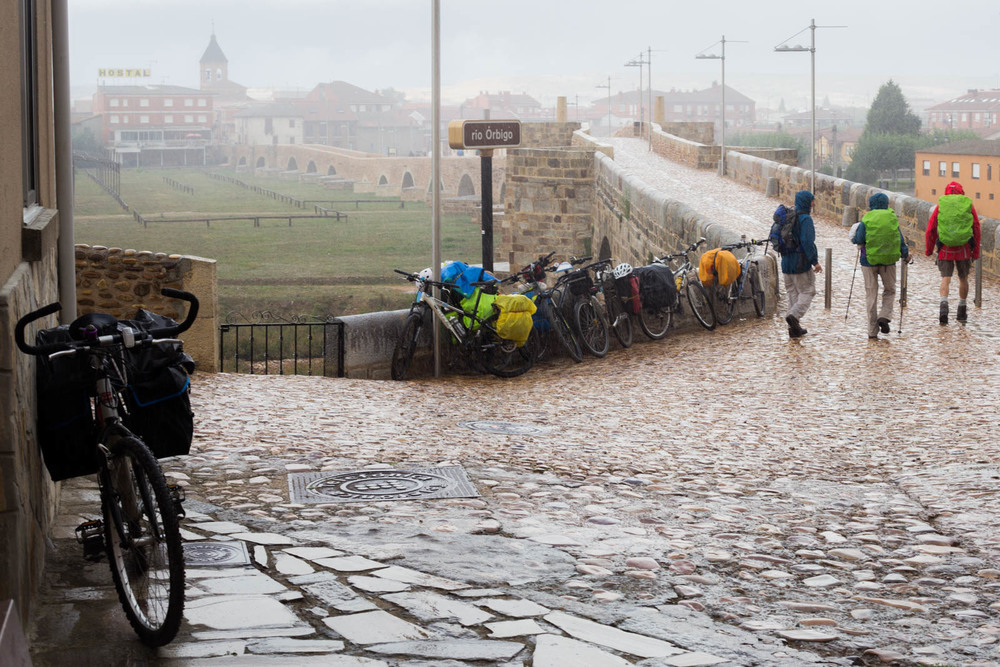 Bicycles with panniers parked along a stone bridge as hikers with backpacks pass by on a rainy day.