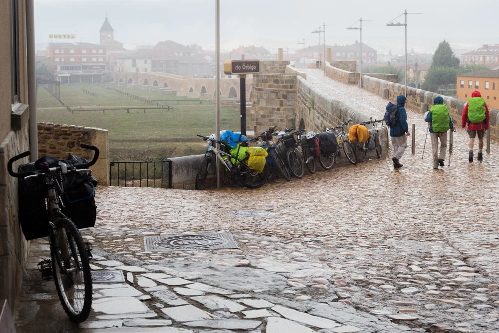 Biciclette con borse laterali parcheggiate lungo un ponte di pietra mentre gli escursionisti con zaini passano in una giornata piovosa.
